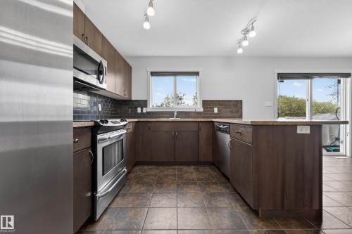 Kitchen featuring dark wood-finish cabinetry, stainless steel appliances, dark tiled backsplash, and stone-look countertops - 5244 1A Avenue, Edmonton, AB 