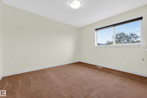 Neutral-toned interior room featuring a large window, brown patterned carpeting, and white baseboards - 5244 1A Avenue, Edmonton, AB 