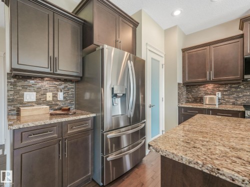Kitchen featuring dark wood cabinetry, stainless steel refrigerator, granite countertops, tile backsplash, and wood-finish flooring - 3 1641 James Mowatt Trail, Edmonton, AB 