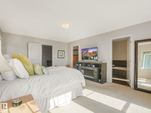 Carpeted sleeping area featuring neutral wall tones, a built-in electric fireplace media console, and a doorway to a tiled bathing area with a soaking tub - 3 1641 James Mowatt Trail, Edmonton, AB 