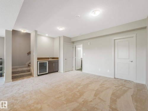 Spacious carpeted room featuring a built-in wet bar with dark wood cabinetry, a beverage refrigerator, and a light-toned countertop - 3 1641 James Mowatt Trail, Edmonton, AB 