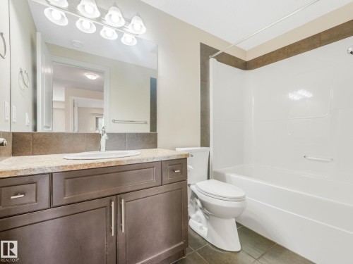 Bathroom featuring a dark wood-finish vanity with a light countertop, an integrated sink, a large wall-mounted mirror, and a white bathtub with a shower - 3 1641 James Mowatt Trail, Edmonton, AB 