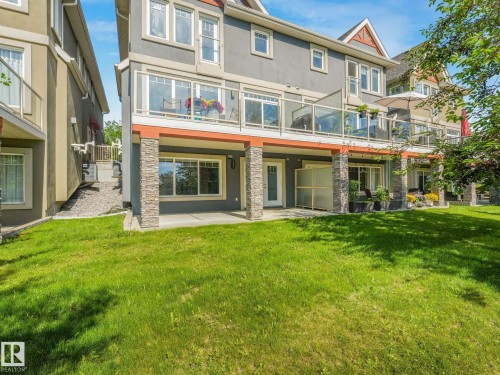 Expansive green lawn backing a multi-story residence featuring a ground-level patio, an upper-level balcony with glass railings, and stone-clad support columns - 3 1641 James Mowatt Trail, Edmonton, AB 