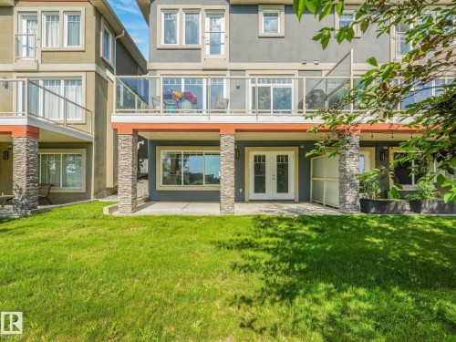 Rear exterior featuring a ground-level patio with double glass doors, stone-clad pillars, a second-story balcony with glass railings, and a manicured lawn - 3 1641 James Mowatt Trail, Edmonton, AB 