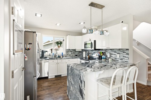 Kitchen featuring white cabinetry, stainless steel appliances, a multi-tone tile backsplash, dark wood-finish flooring, and a large central island with a waterfall countertop - 21384 88 Avenue, Edmonton, AB 