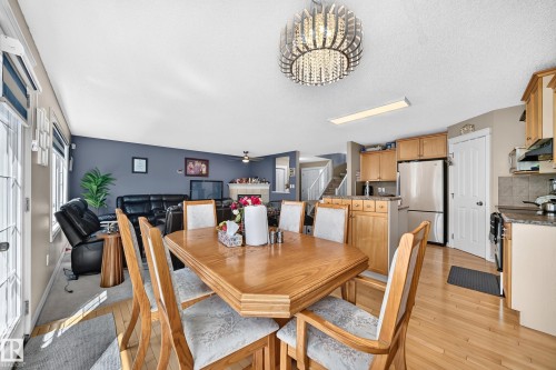 Open-concept living space featuring wood-finish flooring, a contemporary chandelier, a kitchen with light wood cabinetry and stainless steel appliances, and a living area with a fireplace - 3224 26 Street, Edmonton, AB 