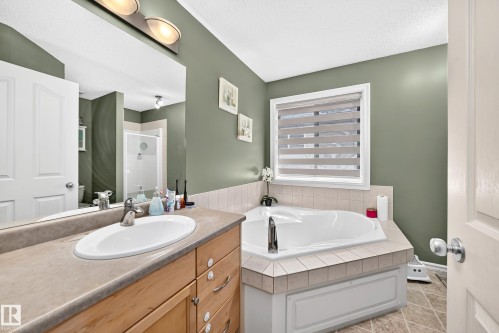 Bathroom featuring an integrated jetted tub with tile surround, a single vanity with a laminate countertop and undermount sink, and a mirrored wall with an overhead light fixture - 3224 26 Street, Edmonton, AB 