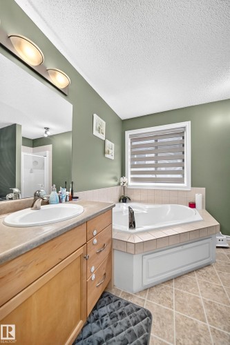 Bathroom featuring a built-in soaking tub with tile surround, a single vanity with an integrated sink and light wood cabinetry, and a window with horizontal blinds - 3224 26 Street, Edmonton, AB 