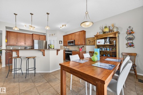Open-concept kitchen and dining area featuring warm wood cabinetry, stainless steel appliances, a breakfast bar with pendant lighting, and tiled flooring - 1310 82 Street, Edmonton, AB 