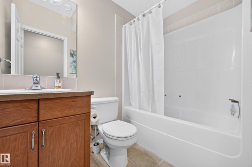 Full bathroom featuring a white tub/shower combination with a tiled surround, a wood-finish vanity with an integrated sink, a large wall mirror, and neutral-toned floor tiling - 1310 82 Street, Edmonton, AB 