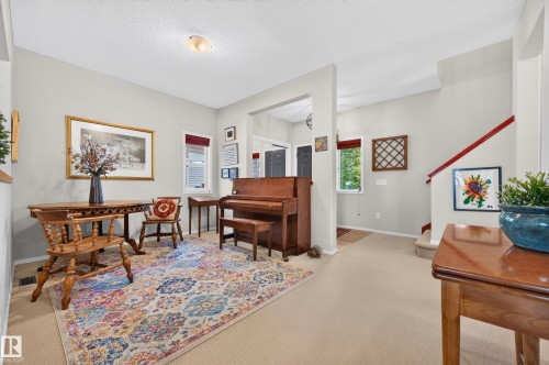 Carpeted living space with light neutral wall paint, featuring a staircase with a red handrail, a front door with dark trim, and multiple windows with red blinds - 1310 82 Street, Edmonton, AB 