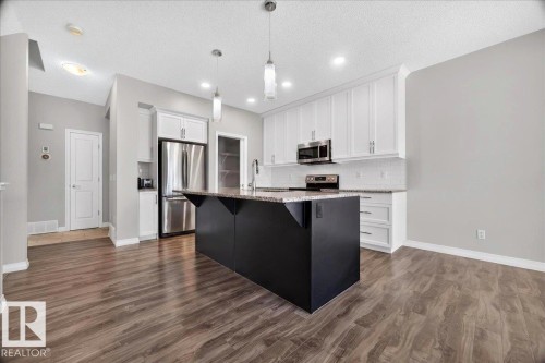 Modern kitchen featuring a large black island with a stone-finish countertop, white upper and lower cabinetry, stainless steel appliances, and wood-finish flooring - 17203 71 Street, Edmonton, AB 