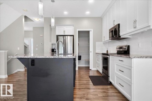 Kitchen featuring a dark-toned island with a double basin sink, white upper and lower cabinetry, stainless steel appliances, and a white subway tile backsplash - 17203 71 Street, Edmonton, AB 