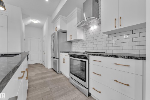 Contemporary kitchen featuring white cabinetry with brushed brass hardware, dark stone countertops, and a white subway tile backsplash - 144 Sunwapta Way, Edmonton, AB 