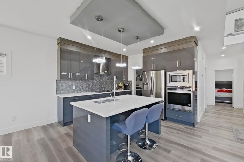 Contemporary kitchen featuring an island with a white countertop and undermount sink, grey high-gloss cabinetry, stainless steel appliances, and a grey hexagon tile backsplash - 415 42 Street, Edmonton, AB 