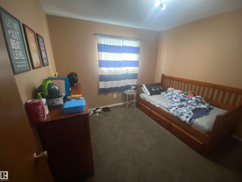 Carpeted interior room with neutral-toned walls, featuring a window with striped curtains, a flush-mount ceiling light fixture, and a built-in wooden daybed with storage drawers - 5713 44A Street, Vegreville, AB 