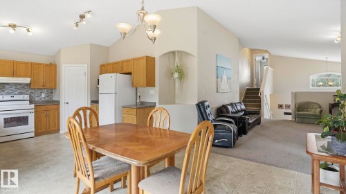 Open-concept living area featuring a kitchen with wood cabinetry and tile flooring, a dining space with a chandelier, and a carpeted living room with a staircase - 6105 52 Avenue, Beaumont, AB 
