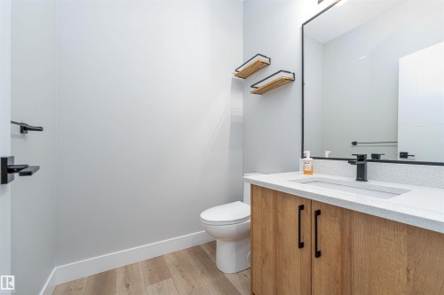 Bathroom featuring a wood-finish vanity with black hardware, a white countertop with an integrated sink, a large framed mirror, and wood-finish flooring - 17320 9 Avenue, Edmonton, AB 