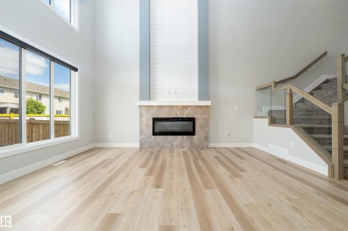 Spacious interior with light wood-finish flooring, a contemporary electric fireplace with a stone surround, and an elegant staircase featuring glass panel railings - 17320 9 Avenue, Edmonton, AB 