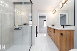 Contemporary bathroom featuring a spacious glass-enclosed shower with light-toned wall tile, a large wood-finish dual vanity with quartz countertops, and matte black hardware - 