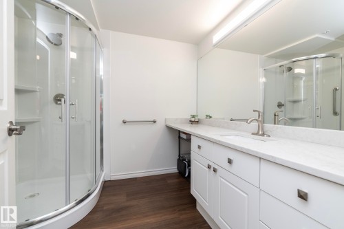 Bathroom featuring a curved glass shower enclosure with built-in shelving, a single vanity with an undermount sink, a light-toned countertop, white cabinetry with square hardware, and wood-finish flooring - 111 182 Haddow Close, Edmonton, AB 