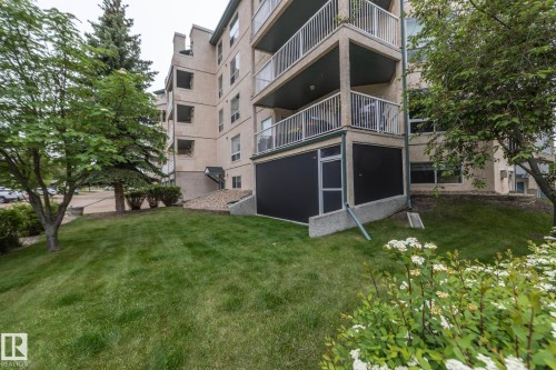 Multi-story building featuring balconies with metal railings, a stucco exterior, and a sloped lawn with mature trees - 111 182 Haddow Close, Edmonton, AB 