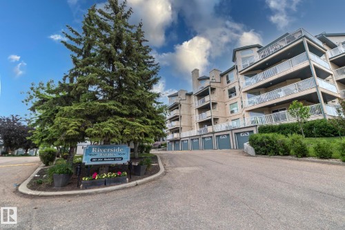 Multi-story residential building featuring private balconies with glass and metal railings, individual garage bays with arched details, and surrounding mature trees and landscaping - 111 182 Haddow Close, Edmonton, AB 