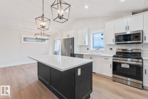 Modern kitchen featuring a large central island with a white countertop, white shaker-style cabinetry, stainless steel appliances, light wood-finish flooring, and contemporary pendant lighting - 301 Fundy Way, Cold Lake, AB - Indoor Photo Showing Kitchen With Upgraded Kitchen