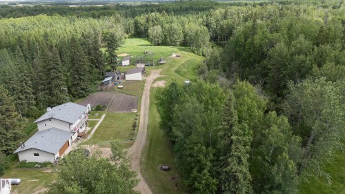 Expansive treed acreage featuring a primary residence with an attached garage, an unpaved driveway, multiple accessory structures, and cultivated garden plots - 58403 Rr 60, Rural Barrhead County, AB - Outdoor With View