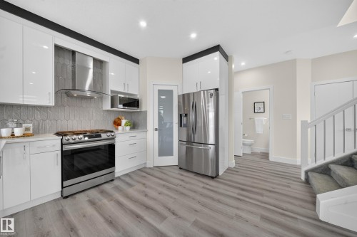 Modern kitchen featuring sleek white cabinetry, stainless steel appliances, a unique geometric tile backsplash, stone countertops, and wood-finish flooring - 1099 Eaton Road, Edmonton, AB 