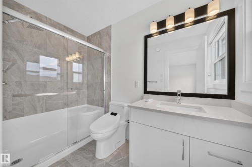 Bathroom featuring a glass-enclosed tub/shower combo with large format tile surround, a built-in vanity with a stone-finish countertop, a single basin sink with a chrome faucet, and a large framed mirror with integrated lighting - 1099 Eaton Road, Edmonton, AB 