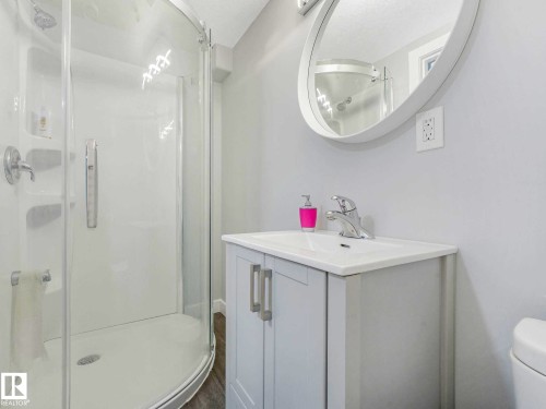 White bathroom featuring a glass-enclosed shower with a curved base, a white vanity with a single basin sink, a circular framed mirror, and wood-finish flooring - 9207 169 Street, Edmonton, AB - Indoor Photo Showing Bathroom