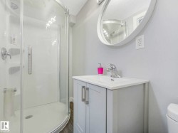 White bathroom featuring a glass-enclosed shower with a curved base, a white vanity with a single basin sink, a circular framed mirror, and wood-finish flooring - 
