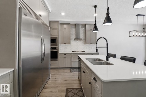 Kitchen featuring a center island with a double basin sink and matte black faucet, stainless steel appliances, light gray cabinetry, white subway tile backsplash, and wood-finish flooring - 3234 Chernowski Way, Edmonton, AB 