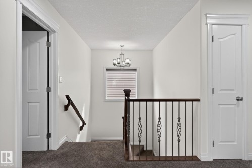 Upper-level landing featuring neutral carpeting, a staircase with dark wood railing and wrought iron balusters, a window with horizontal blinds, and a chandelier with a dark finish - 9628 221 Street, Edmonton, AB 