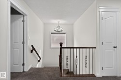 Upper-level landing featuring neutral carpeting, a staircase with dark wood railing and wrought iron balusters, a window with horizontal blinds, and a chandelier with a dark finish - 