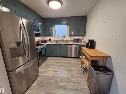 Kitchen featuring sleek teal cabinetry, stainless steel appliances, light-toned wood-finish flooring, a contemporary ceiling light fixture, and a window above the sink - 2514 149 Avenue, Edmonton, AB 