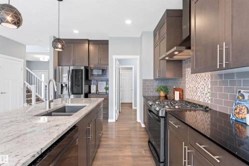 Contemporary kitchen featuring an oversized island with an undermount sink and light-toned stone countertop, dark wood-finish cabinetry, stainless steel appliances, and wood-finish flooring - 9664 223 Street, Edmonton, AB 