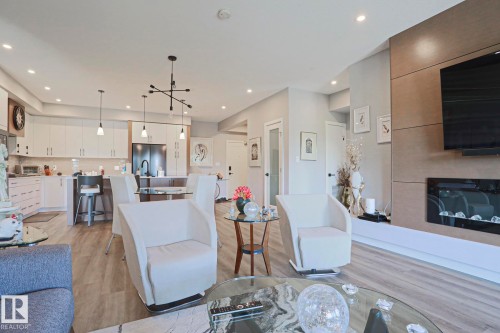 Open-concept living space featuring wood-finish flooring, a contemporary kitchen with white cabinetry, a built-in fireplace, recessed lighting, and modern pendant light fixtures - 211 7463 May Common, Edmonton, AB 