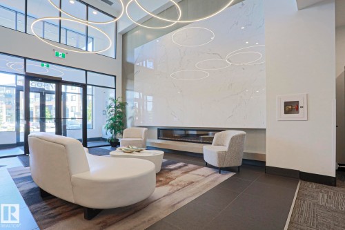 Lobby featuring a grand fireplace with a large-format tile surround, contemporary circular pendant lighting, full-height windows, a glass entry door, and large dark floor tiles - 211 7463 May Common, Edmonton, AB 