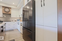 Modern kitchen featuring a black stainless steel refrigerator, white cabinetry with dark hardware, light wood-finish flooring, a contemporary range hood, and a textured tile backsplash - 