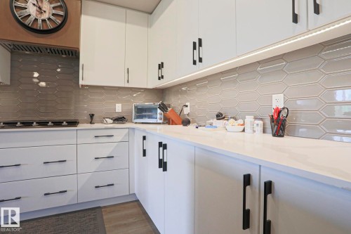 Contemporary kitchen featuring white cabinetry with matte black hardware, light-toned countertops, and a patterned tile backsplash - 211 7463 May Common, Edmonton, AB 
