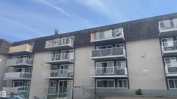 Multi-story apartment building with stucco exterior, individual balconies with white railings, dark grey shingled mansard roof, and multiple large windows - 