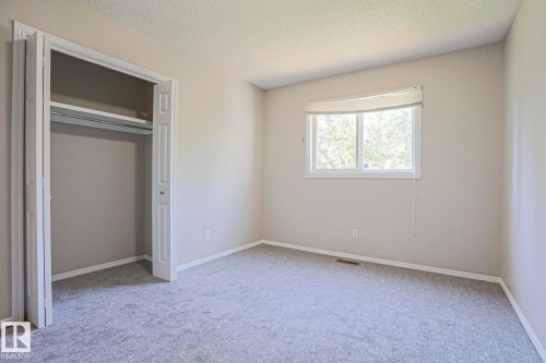 Carpeted room featuring a white-framed window with a roller shade, light-toned walls, a closet with bi-fold doors, and ceiling texture - 17811 62C Avenue, Edmonton, AB 