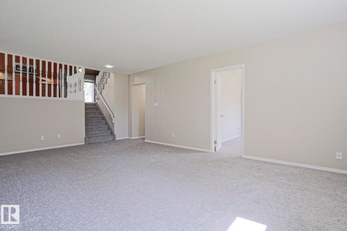 Spacious room featuring light gray carpet, neutral wall paint, white trim, and a visible staircase with white railings - 17811 62C Avenue, Edmonton, AB 