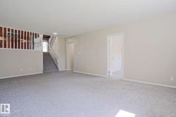 Spacious room featuring light gray carpet, neutral wall paint, white trim, and a visible staircase with white railings - 