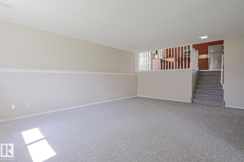 Spacious carpeted room featuring neutral wall tones, white trim, a built-in staircase, and a partial view of an upper-level kitchen with wood cabinetry and a stainless steel refrigerator - 17811 62C Avenue, Edmonton, AB 