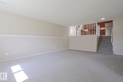 Spacious carpeted room featuring neutral wall tones, white trim, a built-in staircase, and a partial view of an upper-level kitchen with wood cabinetry and a stainless steel refrigerator - 
