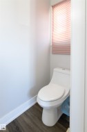 Compact water closet with a white toilet, wood-finish flooring, and a window with blinds - 