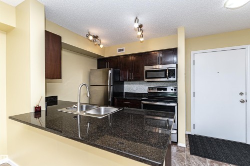 Kitchen featuring a dual basin sink, black countertop, stainless steel appliances, dark wood cabinetry, and track lighting - 404 12045 22 Avenue, Edmonton, AB 